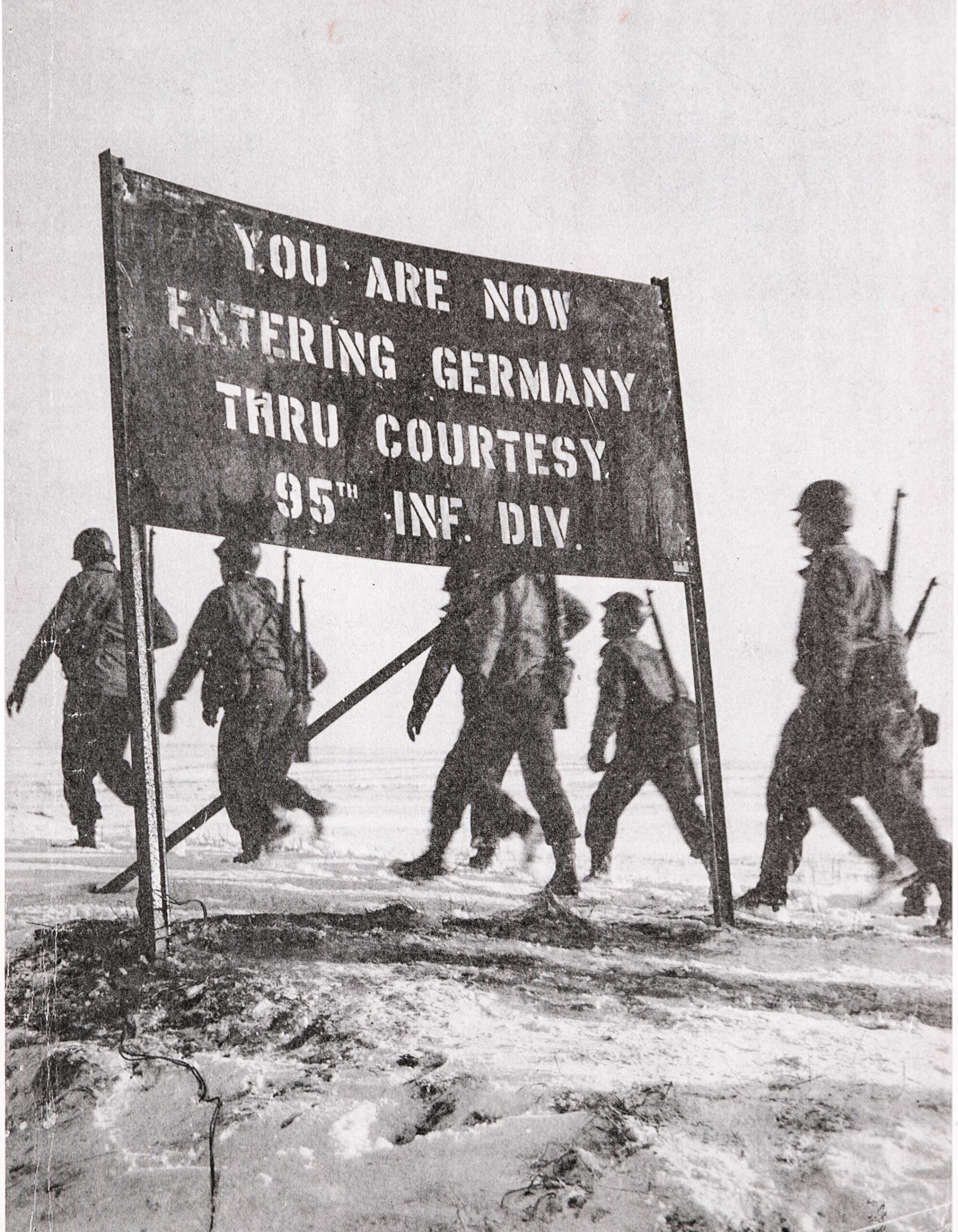 Soldiers walking in field past sign that reads "You are now entering Germany through courtesy 95th Infantry Division"