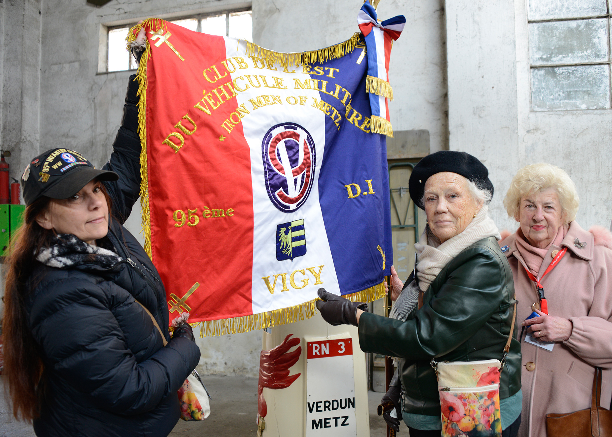 Roselle Calarino and Margaret Jamison (granddaughter and wife of Stephen Jamison, F Co., 378th Reg.) and Irma Madis pose with the new flag in the CEVM motorpool in Vigy.